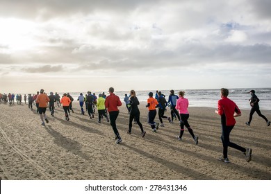 A Group Of Back Lit People In Colorful Outfits Are Running During A Marathon That Goes Partly Over A Beach