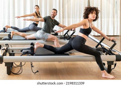 Group of barefoot women and man in activewear spreading arms and looking away while doing stretching exercise on reformers during pilates training in gym