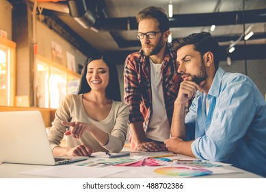 Group of beautiful designers in casual clothes is using a laptop, discussing affairs and smiling while working in studio - Powered by Shutterstock