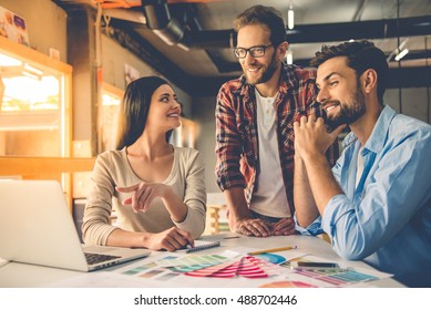 Group of beautiful designers in casual clothes is using a laptop, discussing affairs and smiling while working in studio - Powered by Shutterstock