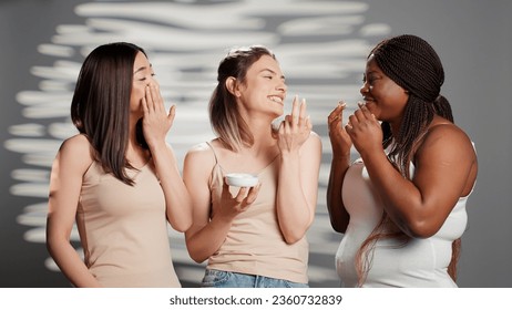 Group of beautiful girls applying moisturizing cream on camera, promoting natural skincare products in studio. Diverse women using moisturizer and serum for new beauty campaign. - Powered by Shutterstock