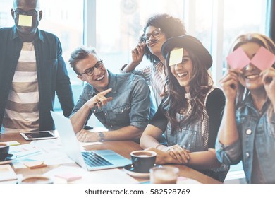 Group of beautiful young people at cafe table with laptop having fun together playing name game with sticky notes to their forehead, laughing