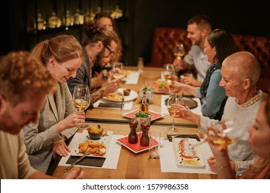 Group Of Best Friends Having Dinner At Restaurant. They All Holding Glasses Of White  Wine, Smiling And Talking.