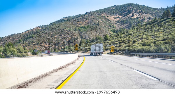 Group of big rigs industrial professional semi\
trucks tractors transporting cargo in different semi trailers\
driving in opposite directions on the divided highway road with a\
long descent signs