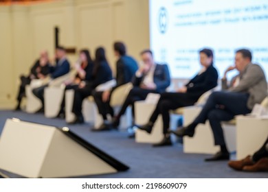 Group Of Blurred Entrepreneurs In Suits Sitting On Chairs Near Large Screen During Business Conference In Modern Auditorium