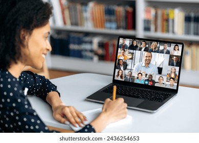 Group brainstorming, online meeting, distance learning. Mixed race female student taking notes in notebook while listening to online lecture on video call,multinational smiling people on laptop screen - Powered by Shutterstock