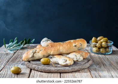 Group of bread sticks stuffed with green olives ion cutting board, olive tree branch, olives in bowl on wooden table with dark background - Powered by Shutterstock