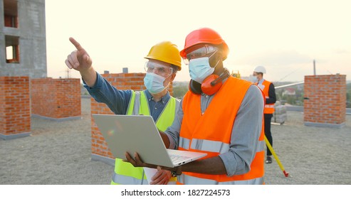 Group of builders in hardhat works on the building site. Two male builders  in medical face mask using laptop computer on construction site. - Powered by Shutterstock