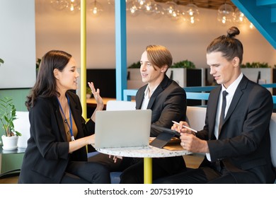 A Group Of Business People, Asian Businesswoman And Caucasian Businessmen, Sitting At A Round Table With Laptop And Tablet, Having A Business Conversation During The Small Conference In A Coffee Shop.