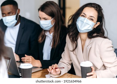 Group Of Business People At Briefing Meeting. Multiracial Coworkers Wearing Medical Masks, Brainstorming Together, Discuss Ideas, Working On New Project, One Woman Looks At Camera