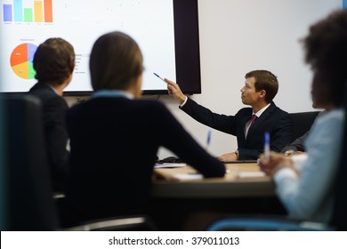 Group Of Business People Meeting In Corporate Conference Room, Smiling During A Presentation. The Coworkers Are Examining Charts And Slides On A Big TV Monitor