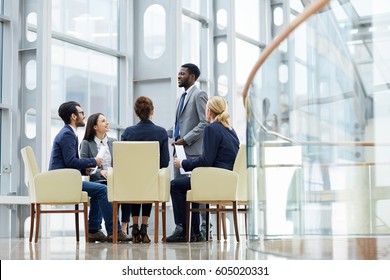 Group Of  Business People Sitting In Circle During Meeting, African  Man Standing Up Giving Speech  In Glass Hall Of Modern Office Building