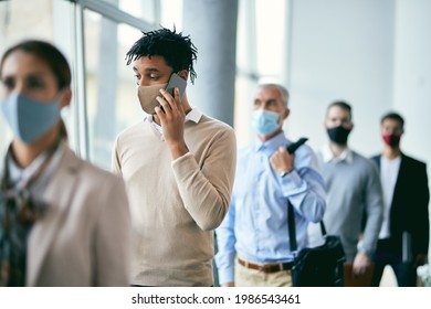 Group Of Business People Standing Six Feet Apart While Waiting For Job Interview In A Hallway. Focus Is On African American Businessman Communicating Over Mobile Phone. 