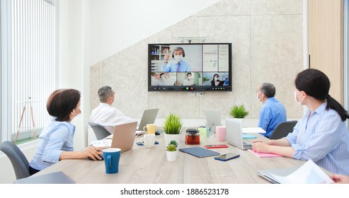 group of business people wearing fack mask are having conference call meeting in boardroom - asian old leader man chatting to colleagues using online video chat on tv screen discussing ideas in office