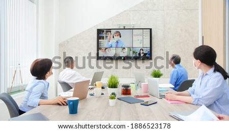 group of business people wearing fack mask are having conference call meeting in boardroom - asian old leader man chatting to colleagues using online video chat on tv screen discussing ideas in office