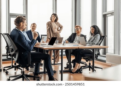 A group of business professionals are having a meeting in a modern office. The meeting is taking place around a large, rectangular table with several laptops and papers on it.