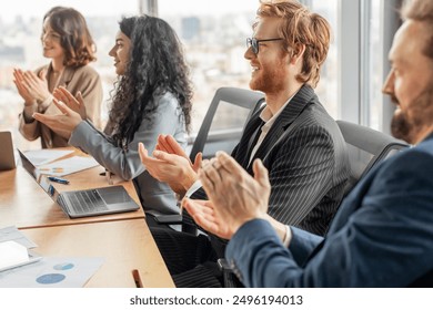 A group of business professionals sit at a table in a modern office, clapping their hands in appreciation as they listen to a presentation. They are dressed in formal attire - Powered by Shutterstock