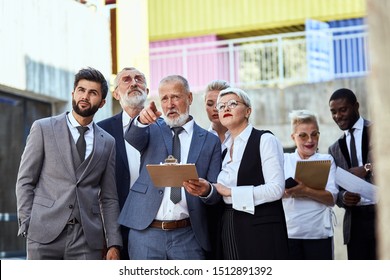 Group Of Businessmen Wear Stylish Suits In The Street. Man In Blue Wear Point Finger Something. Colleagues Near Him Look At It And Listen