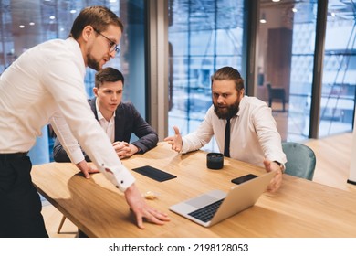 Group Of Businesspeople In Formal Clothes Discussing Details Of Project And Looking At Laptop Screen While Cooperating During Meeting In Modern Boardroom