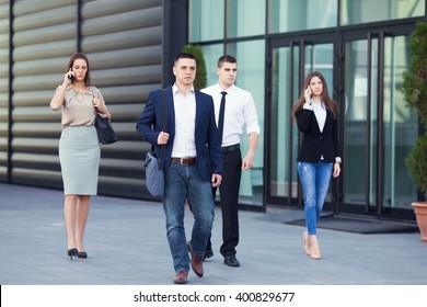 Group Of Busy Business People On The Move In Front Of The Office Building. Focus On Confident Young Man Walking Down The Street