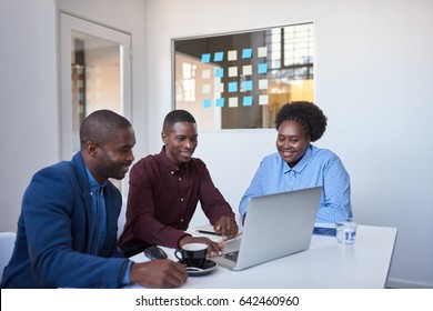 Group Of Casually Dressed African Colleagues Talking Together And Using A Laptop While Sitting At A Table In A Modern Office 