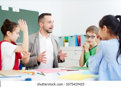 Group Of Caucasian 1st Graders Sitting At Desk With Professional Male Teacher, Drawing And Communicating With Each Other