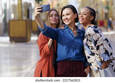 Group Of Cheerful Diverse Female Friends Taking Self Portrait On Cellphone While Standing On Sidewalk Against Blurred Background In City