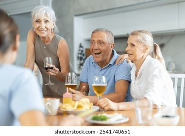 Group of cheerful elderly friends, women and man, gathering around kitchen table, enjoying beer and snacks, engaging in lively conversation and laughter - Powered by Shutterstock