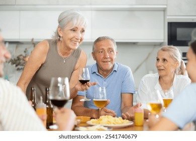 Group of cheerful elderly friends, women and man, gathering around kitchen table, enjoying beer and snacks, engaging in lively conversation and laughter - Powered by Shutterstock