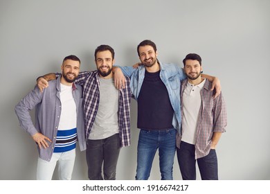 Group Of Cheerful Friends Having Fun Together. Portrait Of Happy Bearded Young Men Standing With Arms Around Each Other's Shoulders And Smiling At Camera In Studio With Light Gray Background