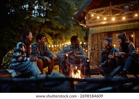 Group of cheerful friends talking while roasting marshmallows over campfire on autumn evening. 