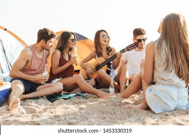 Group Of Cheerful Happy Friends Camping At The Beach, Drinking Beer, Laughing, Playing Guitar