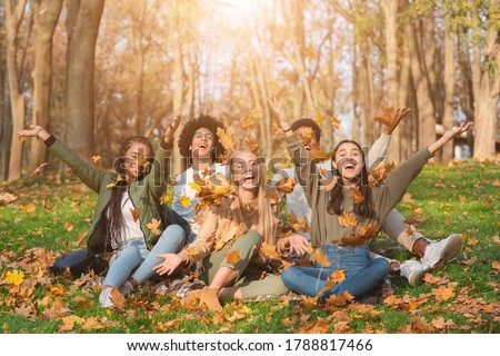 Group of cheerful international friends playing with autumn leaves in public park, taking autumn photos together