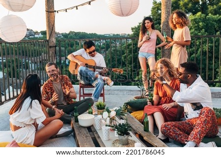 A group of cheerful interracial friends is having a party on the roof. A man on the chair is playing guitar while the rest of the group is chatting, drinking beer, and having fun.