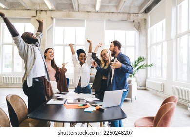 Group of cheerful multiethnic colleagues in casual clothes standing around table with gadgets and raising arms in modern loft office - Powered by Shutterstock