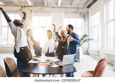 Group of cheerful multiethnic colleagues in casual clothes standing around table with gadgets and raising arms in modern loft office - Powered by Shutterstock