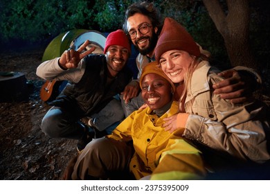 Group of cheerful multi-ethnic friends pose hugging and smiling for outdoor selfie phone. Happy young adults gathered at campsite on winter night. Positive social relations on camping vacation. - Powered by Shutterstock