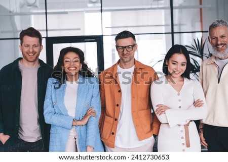Group of cheerful multiracial colleagues standing in modern office near glass wall, looking at camera while male coworkers keeping hands in pockets and smiling young female folding arms across chest