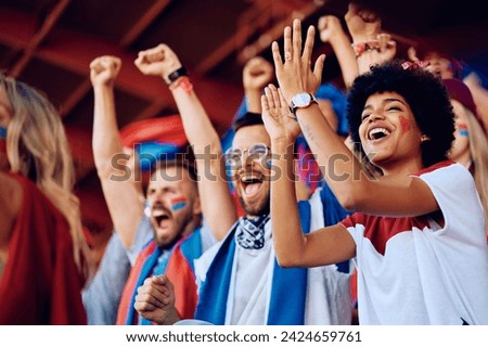 Group of cheerful sports fans celebrating victory of their favorite team while spectating game from stadium stands. Focus is on African American woman.