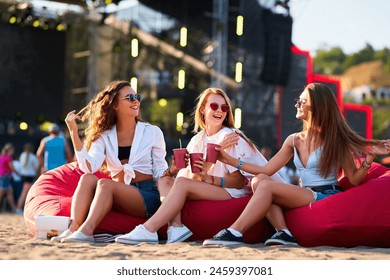 Group of cheerful women lounging on red bean bags, clinking drinks at sunny beach festival. Joyful female friends in casual summer attire enjoy music event, seaside celebration with cold beverages. - Powered by Shutterstock