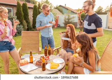 Group of cheerful young friends gathered around the table, eating grilled meat, drinking beer and having fun at a backyard barbecue party - Powered by Shutterstock