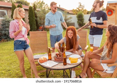 Group of cheerful young friends gathered around the table, eating grilled meat, drinking beer and having fun at backyard barbecue party