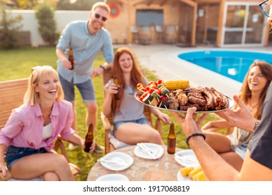 Group Of Cheerful Young Friends Gathered Around The Table, Drinking Beer And Having Fun At Backyard Poolside Barbecue Party While The Host Is Serving Food