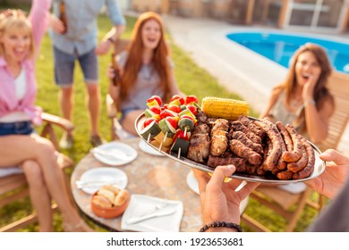 Group Of Cheerful Young Friends Gathered Around The Table, Drinking Beer And Having Fun At A Backyard Poolside Barbecue Party, Host Bringing Plate Of Grilled Meat And Vegetables