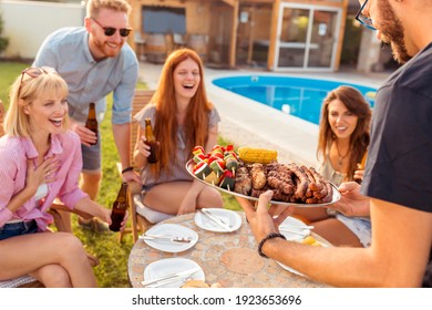 Group Of Cheerful Young Friends Gathered Around The Table, Drinking Beer And Having Fun At A Backyard Poolside Barbecue Party, Host Bringing Plate Of Grilled Meat And Vegetables