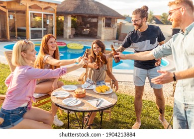 Group of cheerful young friends having fun backyard barbecue party by the swimming pool, making a toast with bottles of beer and enjoying sunny summer days outdoor