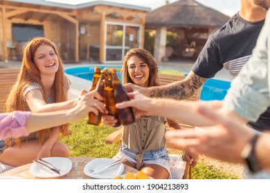 Group Of Cheerful Young Friends Having Fun At Poolside Backyard Barbecue Party, Making A Toast With Bottles Of Beer And Enjoying Sunny Summer Days Outdoor