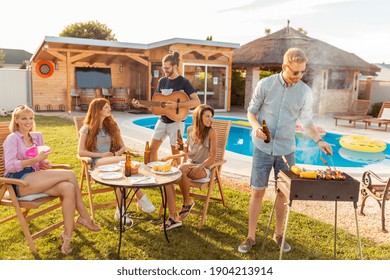 Group Of Cheerful Young Friends Having Fun Playing The Guitar And Singing At Backyard Barbecue Party, Drinking Beer, Grilling Meat And Relaxing By The Swimming Pool