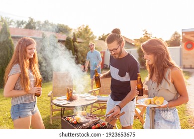 Group Of Cheerful Young Friends Having Fun At Backyard Barbecue Party, Grilling Meat, Drinking Beer, Playing Badminton And Relaxing On A Sunny Summer Day Outdoors