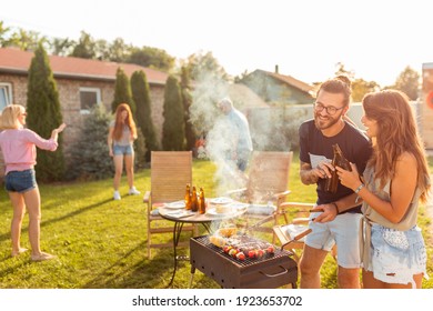 Group Of Cheerful Young Friends Having A Backyard Barbecue Party, Grilling Meat, Making A Toast, Drinking Beer And Having Fun On A Sunny Summer Day